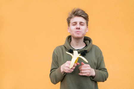 The student enjoys the taste of banana. Young man eats a banana with eyes closed against the background of an orange wall.の写真素材