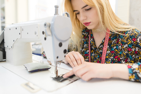 Closeup portrait of a cute young seamstress sitting and sewing on a sewing machine. The seamstress works professionally equipped in the studio.の写真素材