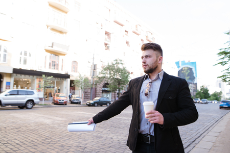 A man with a cup of coffee in his hands stands by the road and catches a taxi. A business man stops a taxi with his hand.の写真素材
