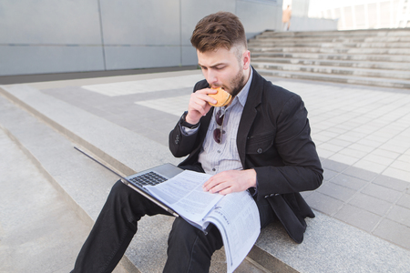 Busy man sits on the ground with a laptop and documents on his lap and snacks. Businessman sitting on the stairs, working on a nude towel and eating.の写真素材