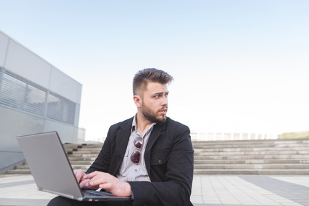 Businessman sits on a staircase with a laptop at the knee-field and works. The banned man works outdoors and looks away. Business concept.の写真素材