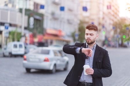 Portrait of a handsome business man standing with a cup of coffee in his hands, looking at the wrist watch against the background of a blurred urban landscape. Busy businessman looks at the clock.の写真素材