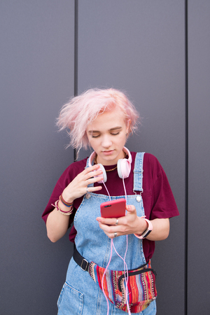 The sweet hipsters girl with stylish clothes, colored hair and headphones is in a dark background and uses a smartphone. Creative girl against the background of a dark wall.の写真素材