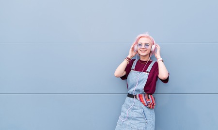Happy, stylish girl with pink hair listening to music in headphones and smiling on a blue background. Smiling girl hipster on the background of a blue wall.の写真素材