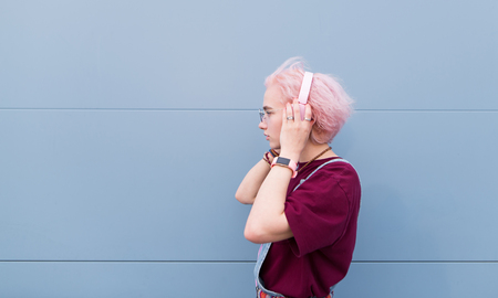 Stylish girl listens to music in pink headphones on a blue background. Portrait of a girl with pink hair and headphones on the background of a blue wall.の写真素材
