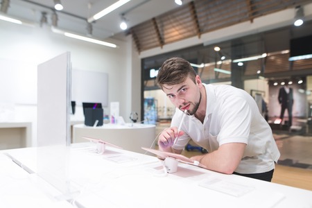 Handsome man stands next to a showcase with tablet stylus in his hands and looks at the camera....