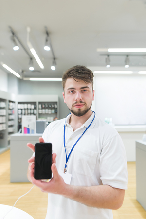 Portrait of a technology store consultant with a smartphone in his hands. The husband's consul is in the electronics store with a smartphone in his hands.の写真素材