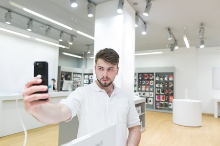 Handsome man makes a selfie on a smartphone in an tech store. Camera test when choosing a smartphone. The buyer selects a smartphone in the electronics store.の写真素材