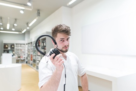 man with a beard and a white T-shirt standing in the electronics store with headphones in his hands and looking into the camera. Buyer with headphones on the background of the technology store.の写真素材