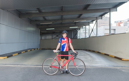 Happy man with a bicycle, cycling clothes stands on the road against the background of architecture and smiles. Portrait of a cyclists man with a red bicycle at the entrance to the car park.の写真素材