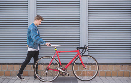 Stylish young man in a denim jacket and a bicycle on a gray background. Portrait of a hystere with a red bicycle on the background of the wallの写真素材