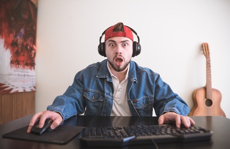 Amazed gamer with beard plays video games at home on a computer. Man with a surprised face sits at the table and plays video games. Portrait of a Surprised Gamer. Video Game Concept.の写真素材