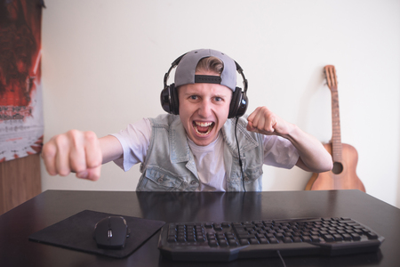 Portrait of a joyful young gamer playing video games at home on a computer. Gamer in headphones and cap is happy because he has won the game. Computer Games Concept.の写真素材