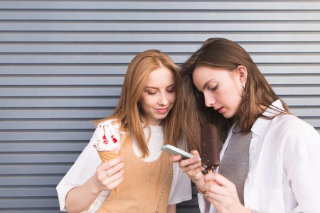 Attractive young girls are standing on the background of a gray wall with a smartphone and ice cream in their hands. Portrait of cute girlfriends on the background wall.の写真素材