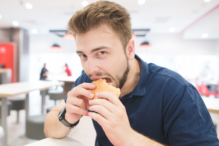 Handsome man sits in a fast food restaurant, bites a burger and looks at the camera. Lunch is a bad fast food. Fastfood concept. student eats a burger in a fast food restaurant.の写真素材