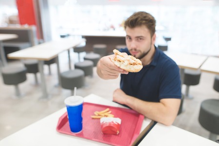 man is sitting at a table in a restaurant with a tray of food and drinks and shows a buggy burger in the camera. A student eats delicious food in the restaurant. Focus on the burger.の写真素材