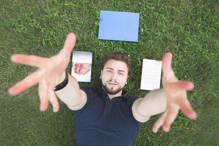 Handsome man in a black T-shirt lays on the grass near the books and extends his hands to the camera. Portrait of a man with a beard lying on the grass, books near the head and looking at the camera.の写真素材