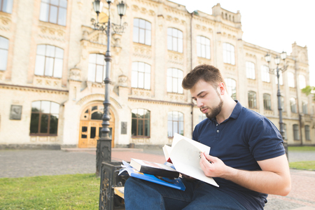 Serious student sitting on a bench at a university campus and studying. Man wearing a beard sitting on a bench on the background of a university building and reading a book.の写真素材