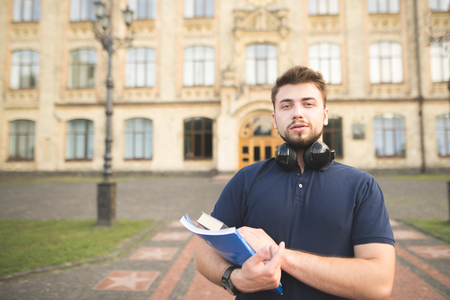 Portrait a man with books in his hands standing on the background of the building and looking into the camera.Student with books in his hands stands on campus at the university and poses to the cameraの写真素材