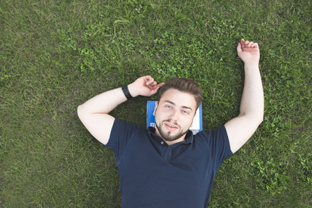 Handsome man with a beard lays on a green lawn on books under his head, looks at the camera and smiles. Happy student lays a green grass books under his head. Rest from studyingの写真素材