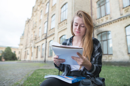 Serious girl sits on a bench on a campus with a book in her arms and reads. Student is sitting at the bench near the university and doing homework. Student Conceptの写真素材