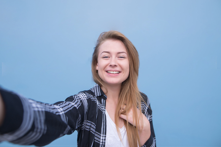 Portrait of a girl standing on a blue background and making a portrait on the camera. Happy girl takes a selfie on a bright blue background.の写真素材