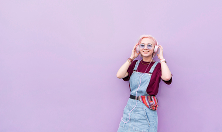 Happy, stylish girl with pink hair listening to music in headphones and smiling on a purple background. Smiling girl hipster on the background of a purple wallの写真素材