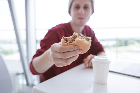 Young man sits in a bright fast food restaurant and holds a burger in his hands. Burger in the hands of a young man close-up and focus. Student dishes fast food.の写真素材