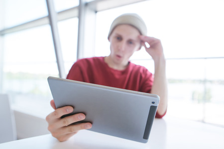 Young man sitting at a table near a window with a tablet in his hands. Young businessman in casual clothing, sitting in a cafe and using a tablet. Tablet close-up and focusの写真素材