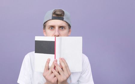 Funny young man with astonished gaze hid behind a book on the background of a purple wall. Portrait of a surprised, funny student with a book on a pastel purple backgroundの写真素材