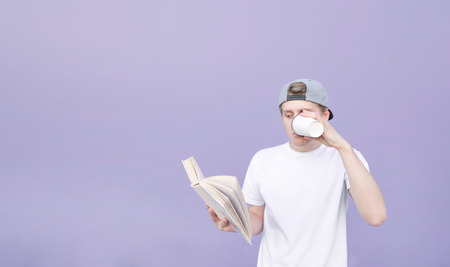 Portrait of a young man in a cap and a white T-shirt who reads a book and drinks coffee on a pastel purple background. Student reads a book and drinks coffee on the background of a purple wall.の写真素材