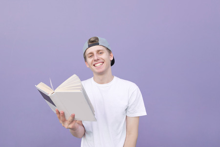 Portrait of a smiling student in a white T-shirt standing on a purple background with a book in hands, smiling and looking at the camera. Happy young man with a book on the background of a purple wallの写真素材