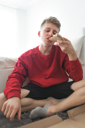 Attractive young man in a red sweatshirt sits in his room on the floor, eats a pizza from the box and gets pleasure. For lunch pizza delivery. Teen eating delicious pizza at home in weekendの写真素材