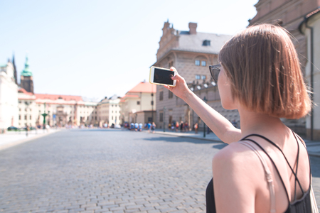 Woman in a black dress walks on a sunny summer day along the streets of the old town and makes a photo on a smartphone. Tourist girl photographs the streets of Prague, Czech Republicの写真素材