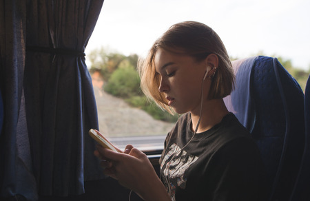 Attractive girl sits in a bus near the window, listens to music and uses a smartphone.Girl travels...