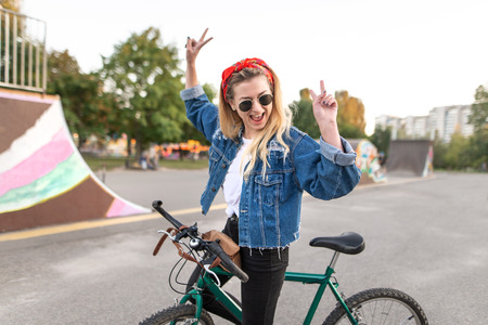Happy young woman stands with a bicycle on the background of a skate park and rejoices with raised hands. Joyful hipster girl riding a bicycle in the city.の写真素材