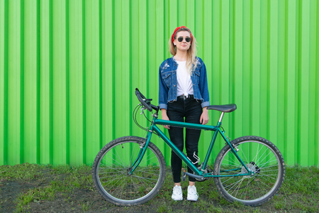 Portrait of a stylish young woman at full height, standing with a bicycle on the background of the wall and looking into the camera. Fashionable woman with a bicycle on a green background. Copyspaceの写真素材