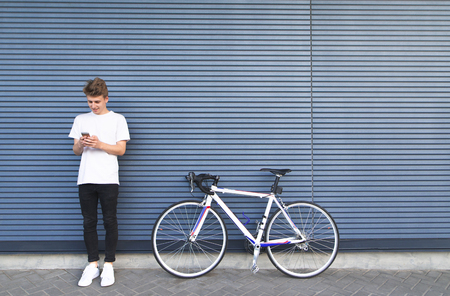 Young man in a white shirt standing near the road bike and uses the smartphone. Happy student standing by a bicycle on the background of the wall.の写真素材