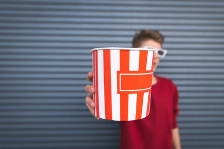 Guy stands on a dark background, holds a bowl of popcorn in his hands and shows him in the camera. Cup with popcorn in the hands of a man close-up.の写真素材