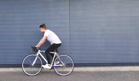 Young man in a white T-shirt rides on a white highway on the background of the wall. Portrait of a student riding a bicycle on the background of the wall.の写真素材