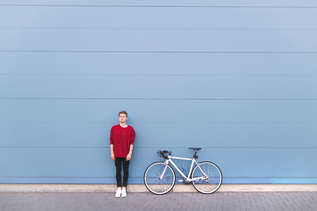 Minimalist portrait of a young man standing with a bike on a pastel blue background and looking into the camera. Stylish man with a bike. Sports concept.Wall. Copyspaceの写真素材