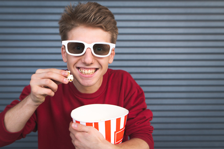Happy young man in 3D eyepieces eats popcorn from a bowl, looks into the camera and smiles, isolated on a dark background.Concept.Copyspaceの写真素材