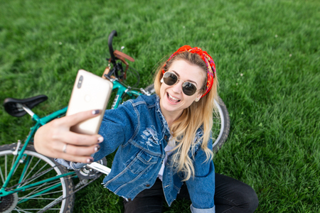 Portrait of an attractive girl sitting on a green grass in a bike and takes selfie on a smartphone. Smiling stylish woman posing for the camera smartphone outdoors in the park.の写真素材