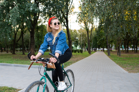 Stylish, happy girl in stylish clothes and sunglasses rides a bike in the park, looks sideways and smiles. Walk on the bike in the park. Active in the rest of the concept.の写真素材