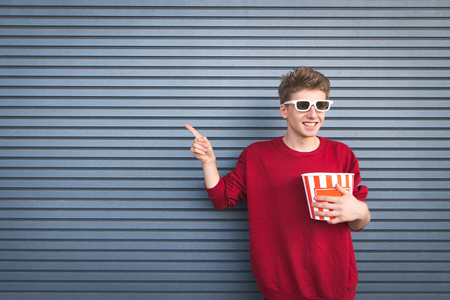 Happy young man in red sweatshirt and white 3D glasses standing with a cup of popcorn in his hands and showing his hand to the side copyspÐ°ce. Background is a dark wall.Cinema Concept.の写真素材