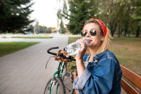Portrait of a stylish girl in social glasses sitting on a bench in the park, resting and drinking water from a bottle. Cyclist girl sits on the bench and drinks water. Activities outdoorsの写真素材