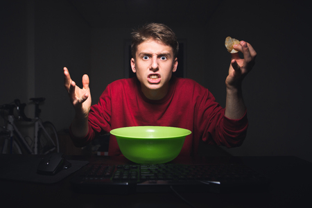 Young boy with the chips in his hand is looking at the computer screen with an outrage. Teenager is looking angrily at the monitor while watching videos on the Internet and eating snacks.の写真素材