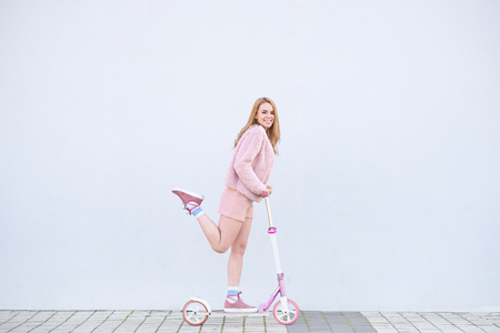 Happy girl in pink clothes rides on a pink kick scooter on the background of a white wall, smiles and looks at the camera. Copyspaceの写真素材