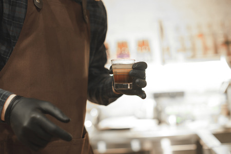 Barista holding a glass with coffee in a coffee shop. Close up photo of the coffee shop owner holding a glass with espresso at the cafe. Focus on a glass. Background photo.の写真素材