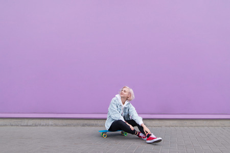 Stylish happy girl sits on a skateboard against a purple wall and smiles. Portrait of a pretty blond...